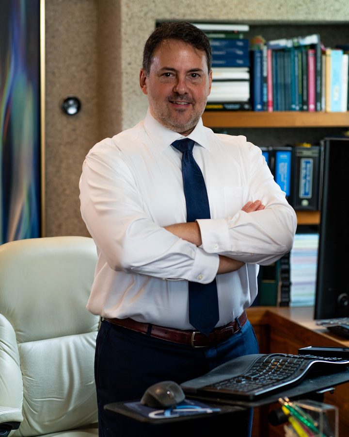 Akiva Shapiro, standing behind his desk with arms crossed, in a white shirt and navy tie, bookshelves behind him