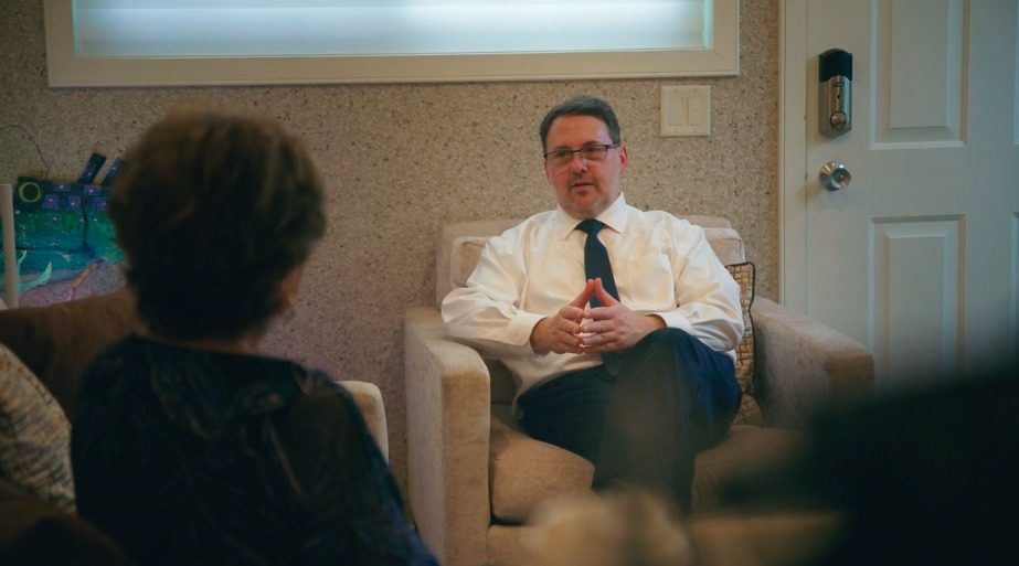Akiva Shapiro, seated in an armchair in a white shirt and tie, in conversation with a client seen from behind