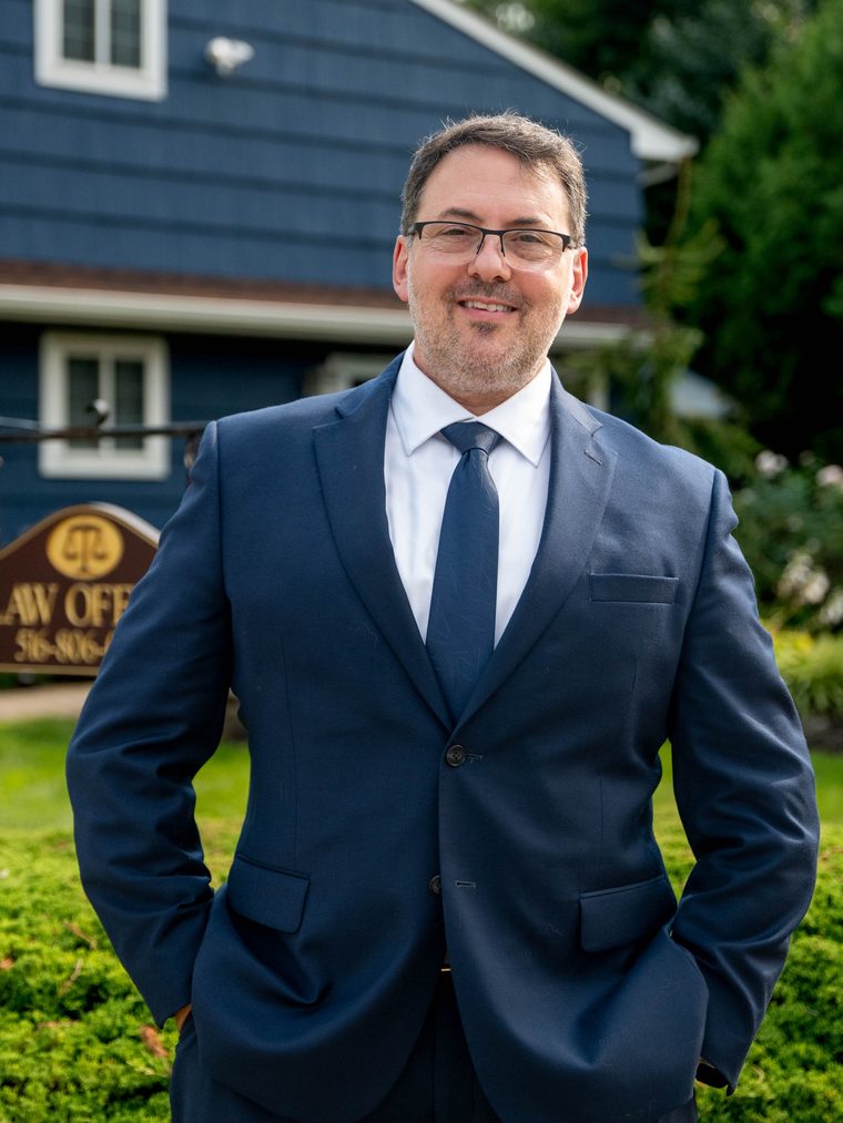 Akiva Shapiro, Esq., smiling, standing outside the firm's office beside its sign